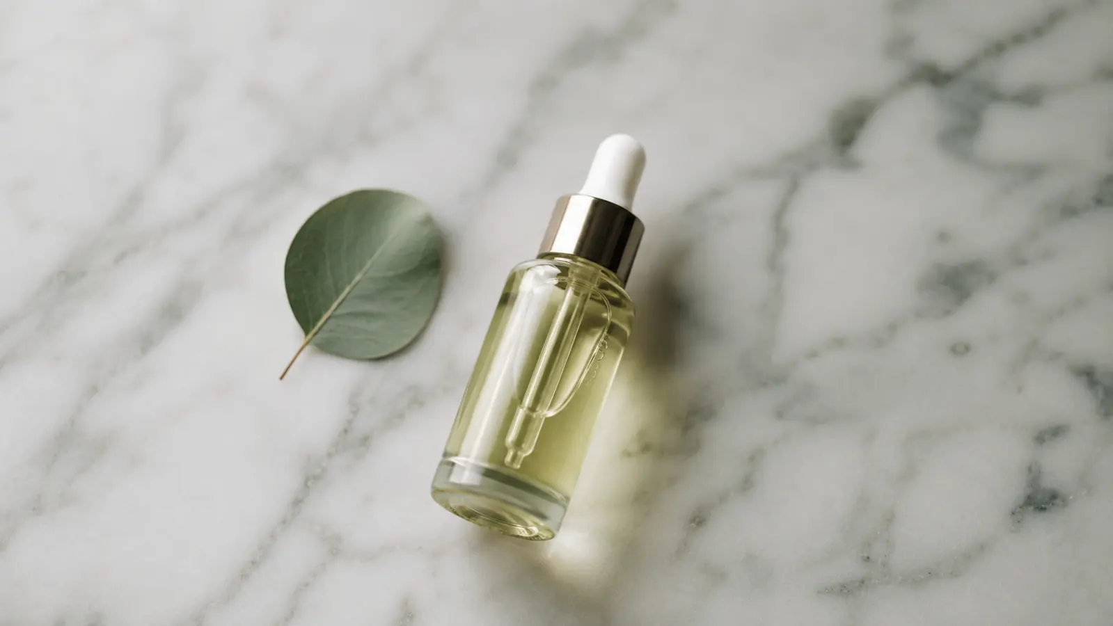 Small dropper bottle of niacinamide serum resting on a white marble surface next to a single eucalyptus sprig
