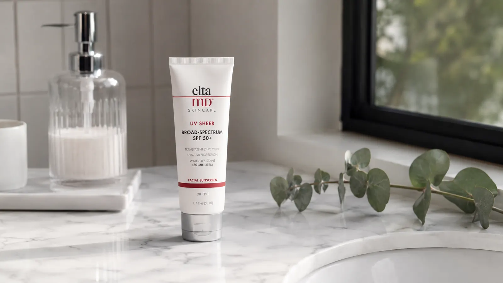 White EltaMD UV Sheer SPF 50 tube on a marble bathroom shelf beside a glass of water and a small plant