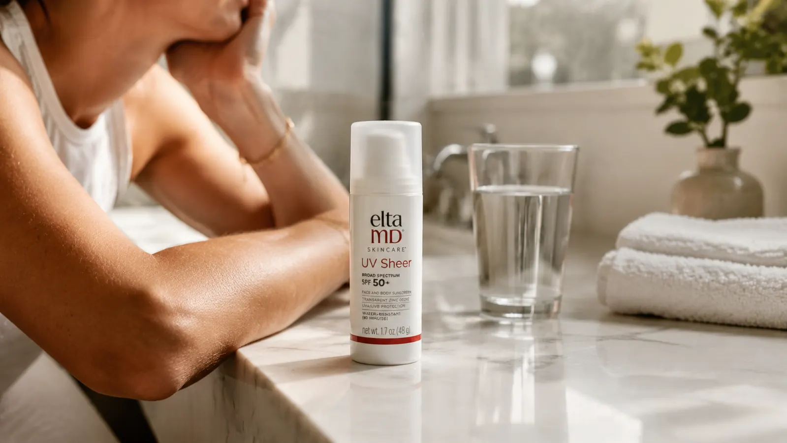 EltaMD UV Sheer SPF 50 bottle on a marble counter next to a glass of water and a folded white towel in soft morning light