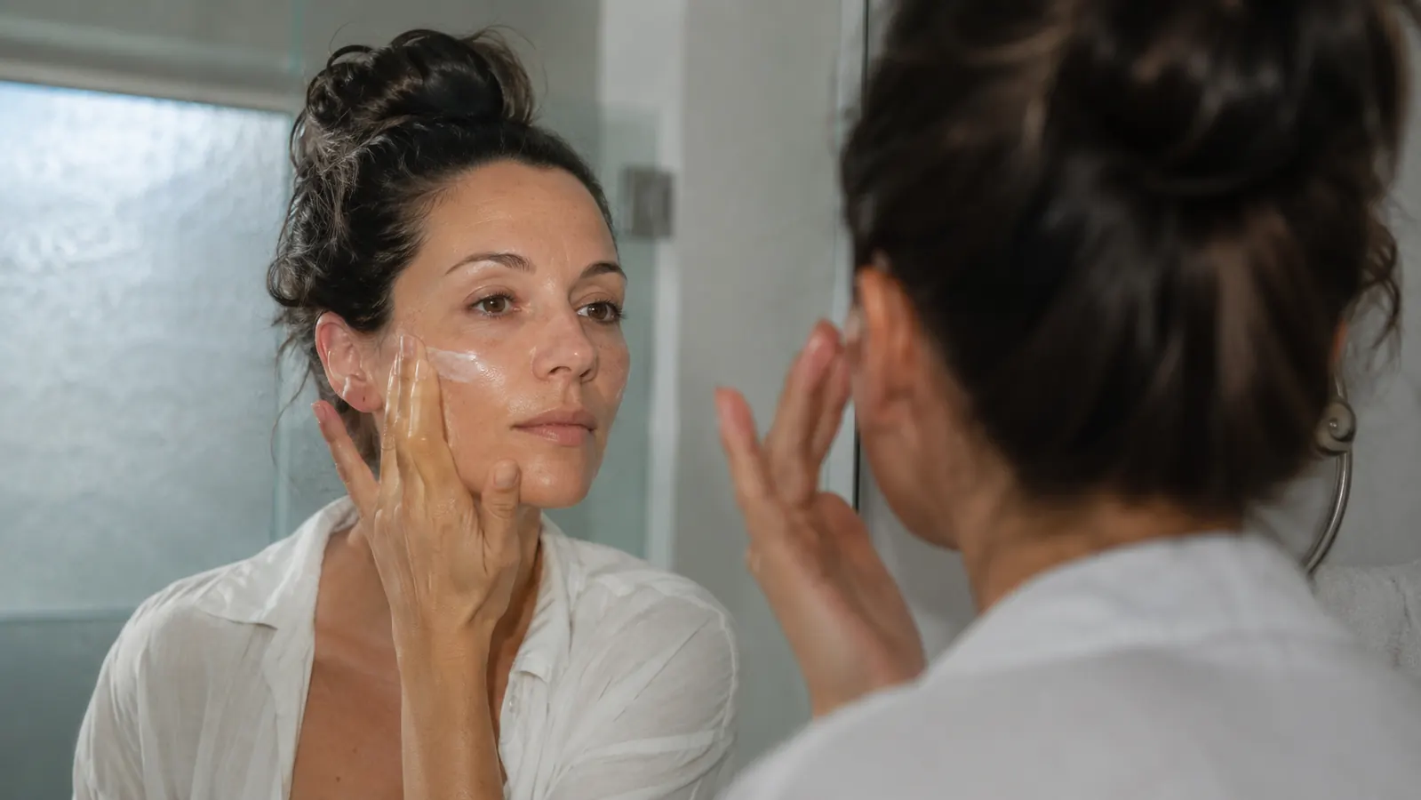 Woman applying sunscreen to her face at a bathroom mirror in morning light