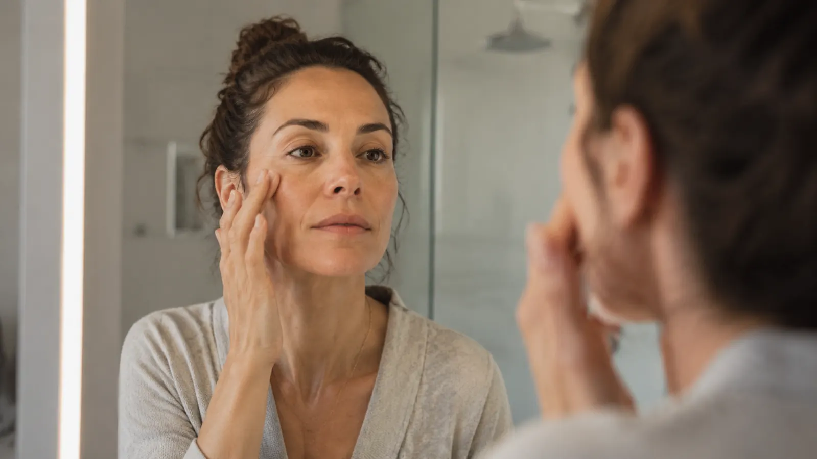 Woman with clear, calm skin applying a small amount of serum to her cheekbone in a well-lit bathroom mirror